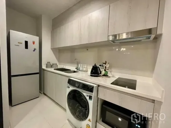 Condominium for rent in Pathum Wan, Bangkok - Modern galley kitchen with white cabinetry, cooktop, fridge and washer-dryer.
