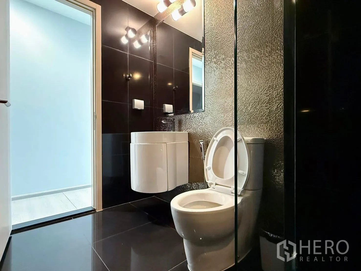 Condominium for rent in Khlong Toei, Bangkok - Modern black-tiled bathroom with wall-mounted sink, toilet, and mirror lights.