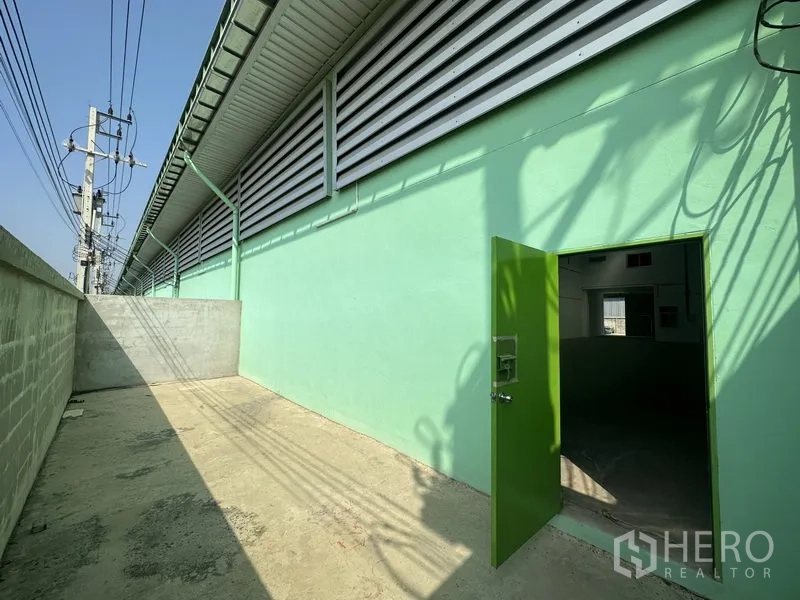 Warehouse for rent in Pathum Thani - Secure side yard with green service door and ventilation louvres along the wall.