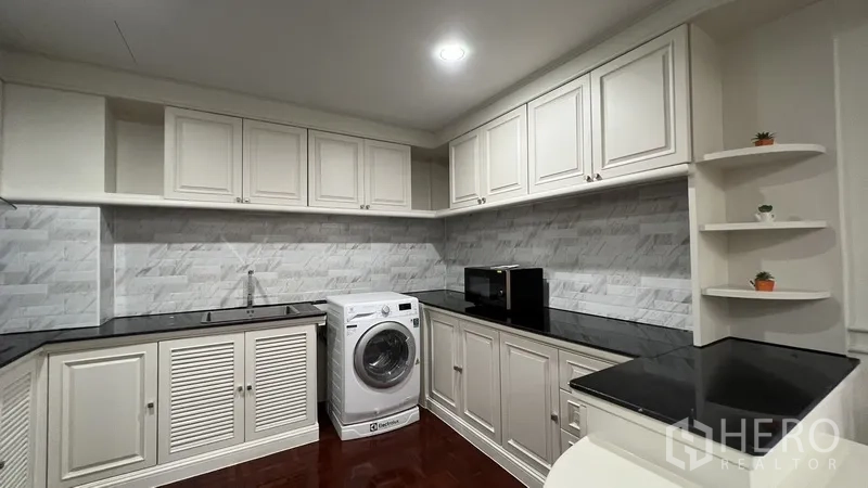Apartment for rent in Watthana, Bangkok - U-shaped kitchen with white cabinetry, black countertops, marble-look tiles, washer and microwave.