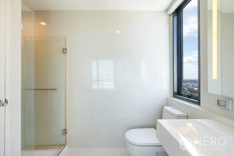 公寓 出租在Watthana, Bangkok - 现代浴室带玻璃淋浴房、采光窗与白色洗手台，远眺城市。