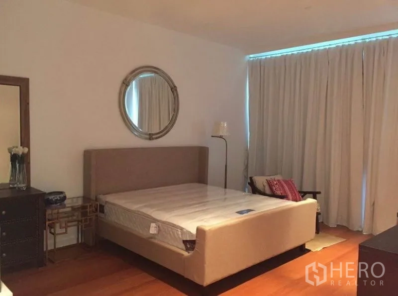 公寓 出租在Pathum Wan, Bangkok - 主卧配软包床、圆形墙镜、窗帘与木地板，灯光柔和。