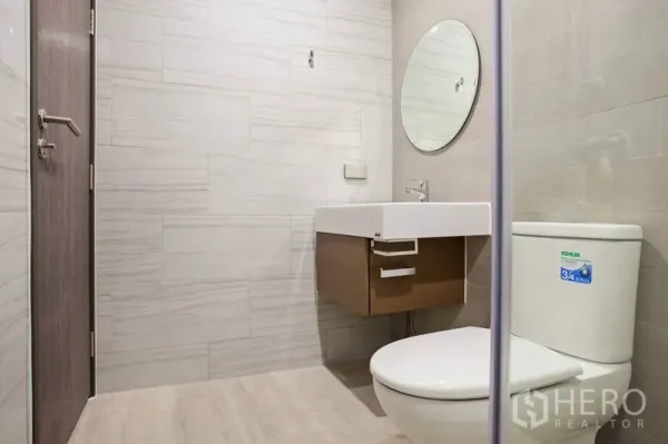 Condominium for rent or sale in Watthana, Bangkok - Contemporary bathroom with wall-mounted vanity, round mirror and Kohler toilet in neutral tones.