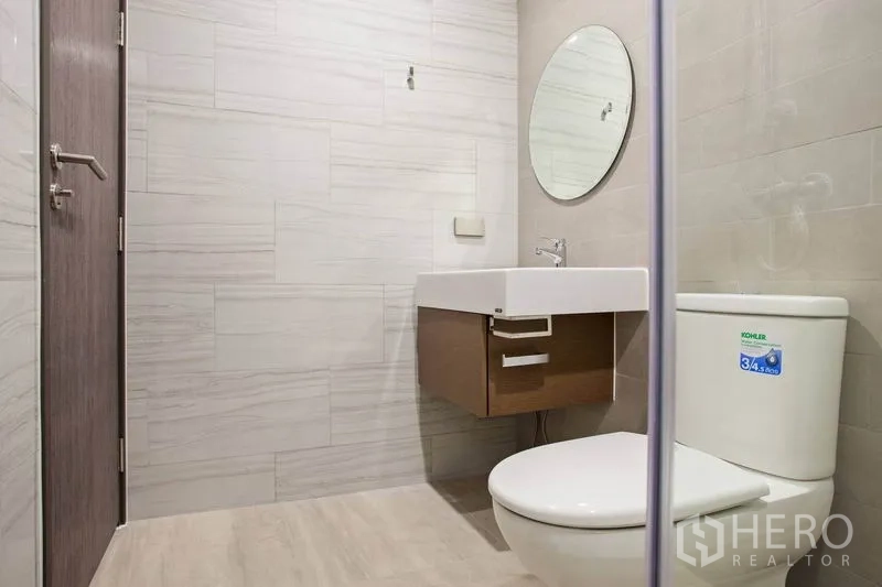 Condominium for rent or sale in Watthana, Bangkok - Contemporary bathroom with wall-mounted vanity, round mirror and Kohler toilet in neutral tones.