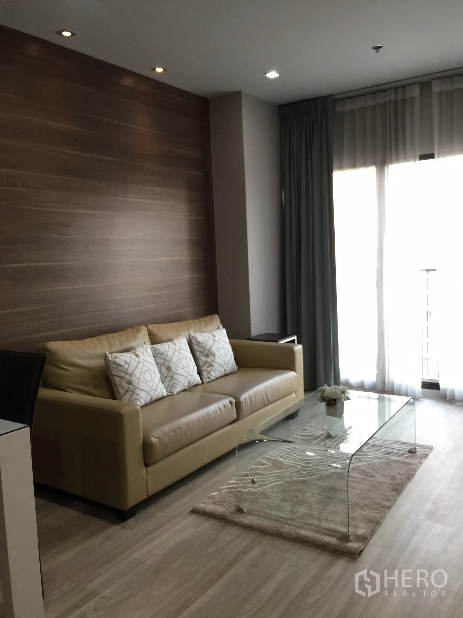 คอนโด ให้เช่าหรือขายในBangkok - ห้องนั่งเล่นโซฟาหนังสีน้ำตาลอ่อน โต๊ะกลางกระจก และแสงจากระเบียง