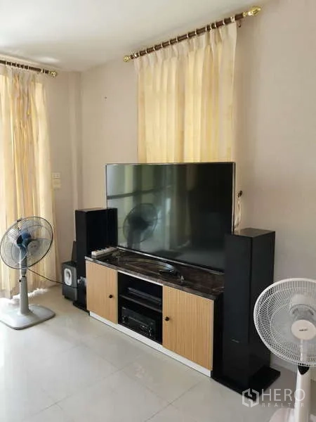 บ้าน ให้เช่า - มุมบันเทิงทีวีจอใหญ่บนตู้ พร้อมลำโพงและพัดลม ตั้งอยู่ในห้องสว่างพื้นกระเบื้อง