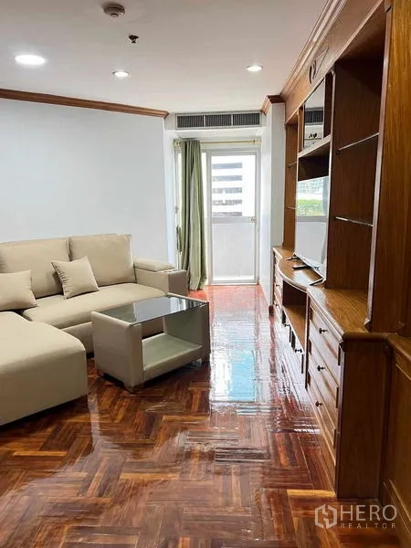 Condominium for rent in Ratchathewi, Bangkok - Living area with built-in wooden TV cabinet, sectional sofa and access to a small balcony.