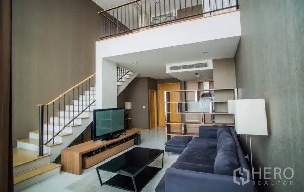 Condominium for rent or sale in Khlong Toei, Bangkok - Double-height living room with staircase, sectional sofa, and TV beneath mezzanine.