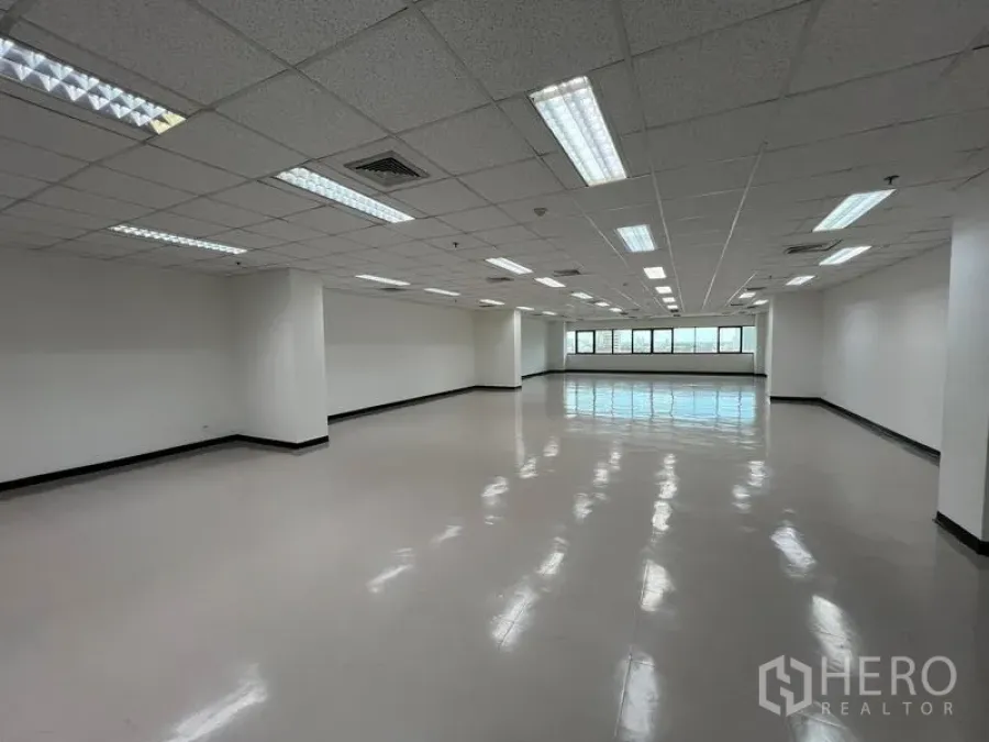 Office Space for rent or sale - Spacious empty office with fluorescent lighting, support columns, and city-view windows at the far end.