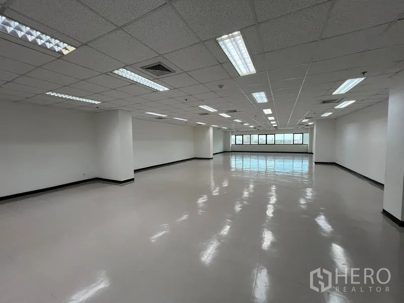 Office Space for rent or sale - Spacious empty office with fluorescent lighting, support columns, and city-view windows at the far end.