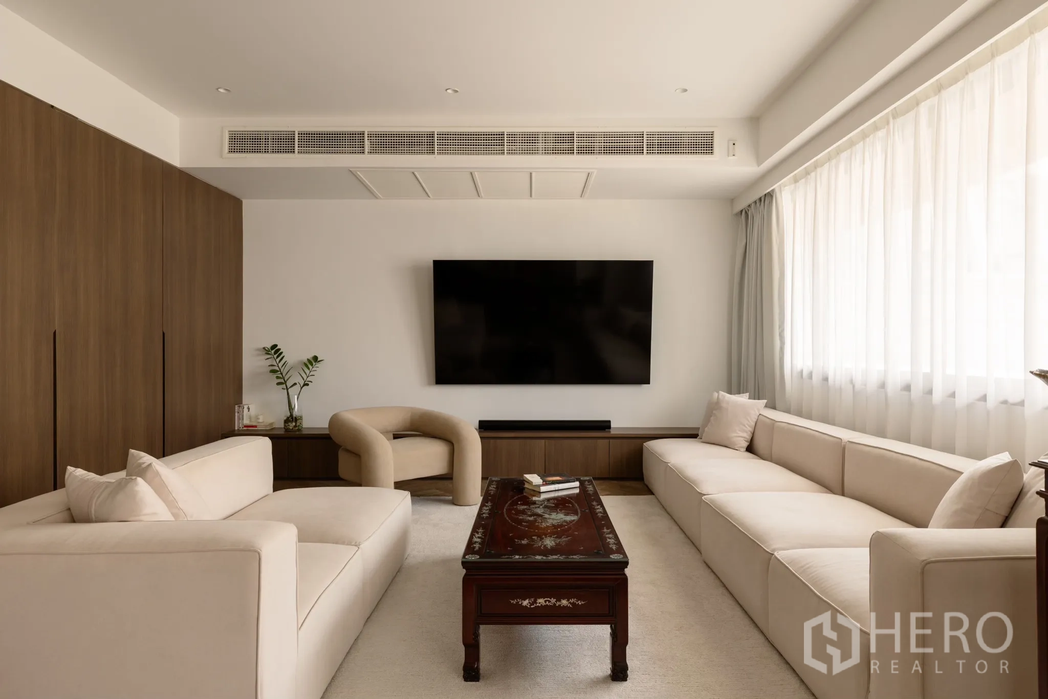 Condominium for sale in Khlong Toei, Bangkok - Bright living room with cream sofas, wall‑mounted TV, wood media console and sheer curtains.