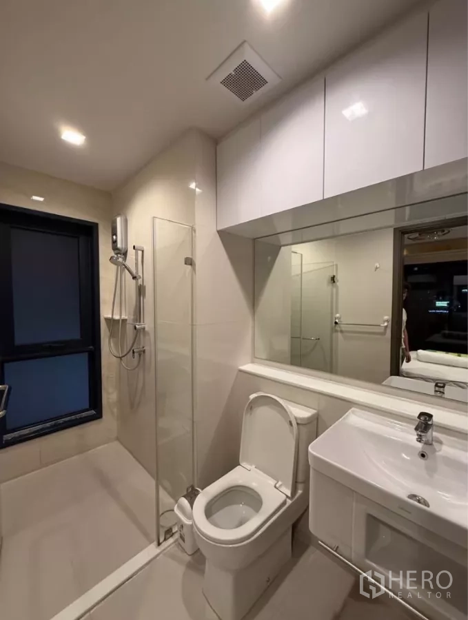 Condominium for rent in Chatuchak, Bangkok - Contemporary bathroom with glass-enclosed shower, vanity, large mirror, and window ventilation.