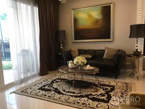 Townhouse for rent or sale in Bang Kho Laem, Bangkok - Elegant living room with leather sofa, abstract artwork, coffee table and patterned rug beside floor‑to‑ceiling sliding glass doors.