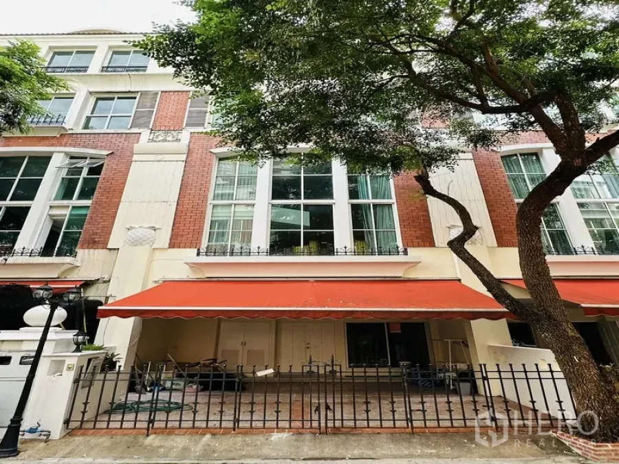 Townhouse for rent in Watthana, Bangkok - Four-storey townhouse facade with red brick accents, tall windows, orange awning and a gated patio beneath a mature tree.