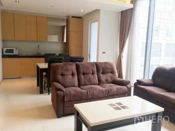 Condominium for rent in Bang Rak, Bangkok - Open-plan living room with brown sofas, dining table, and light wood kitchen cabinetry.