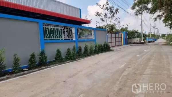 Warehouse for rent in Min Buri, Bangkok - Side exterior of the warehouse showing long boundary wall with metal windows, shrubs and quiet street with power lines.