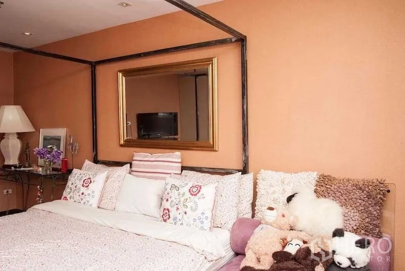 Condominium for rent in Watthana, Bangkok - Cozy bedroom with canopy-style bed, floral pillows, gold-framed mirror and warm peach walls.