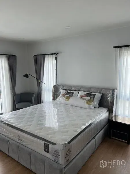 Detached House for rent in Bang Phli, Samut Prakan - Bright master bedroom with upholstered bed, corner chair and dual aspect windows with curtains.