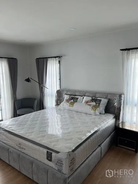 Detached House for rent in Bang Phli, Samut Prakan - Bright master bedroom with upholstered bed, corner chair and dual aspect windows with curtains.