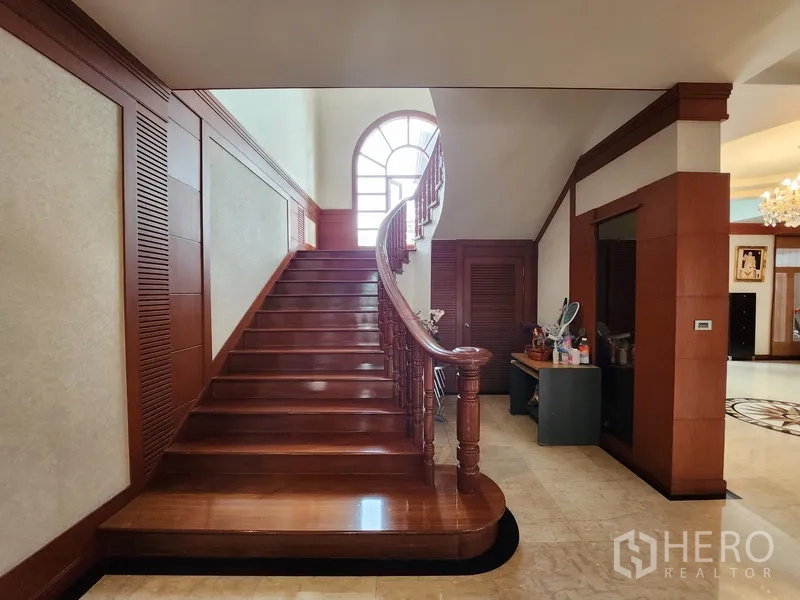 独立式住宅 出租在Prawet, Bangkok - 近景展示宽阔的实木楼梯，配经典栏杆与拱形窗。