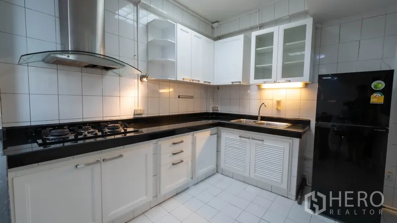 Townhouse for rent in Khlong Toei, Bangkok - White L‑shaped kitchen with black countertops and double‑burner gas stove.