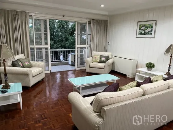 房间 出租在Watthana, Bangkok - 客厅通向阳台，通过玻璃门可见绿树景观。