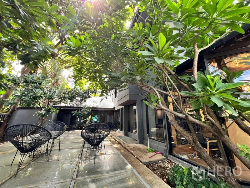 零售空间 出租在Bangkok - 树荫下的户外露台，黑色金属网椅与鸡蛋花树。