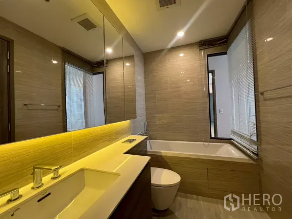 Condominium for rent in Phra Khanong, Bangkok - Modern bathroom with long backlit vanity, bathtub, and neutral wall tiles.