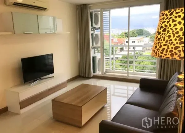 Condominium for rent in Phra Khanong, Bangkok - Living room with brown leather sofa, coffee table and TV beside sliding glass doors to a balcony with leafy neighborhood views.