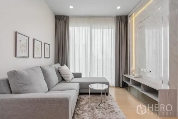 Condominium for sale in Yan Nawa, Bangkok - Modern lounge with L-shaped grey sofa, plush rug and TV feature wall with ambient lighting.