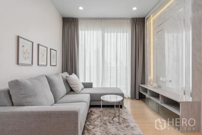 Condominium for sale in Yan Nawa, Bangkok - Modern lounge with L-shaped grey sofa, plush rug and TV feature wall with ambient lighting.