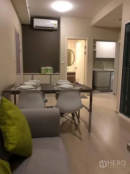 Condominium for rent or sale in Huai Khwang, Bangkok - Modern dining and living area in a Bangkok condo with a set dining table, white chairs, gray sofa, and view toward the kitchen and bathroom.