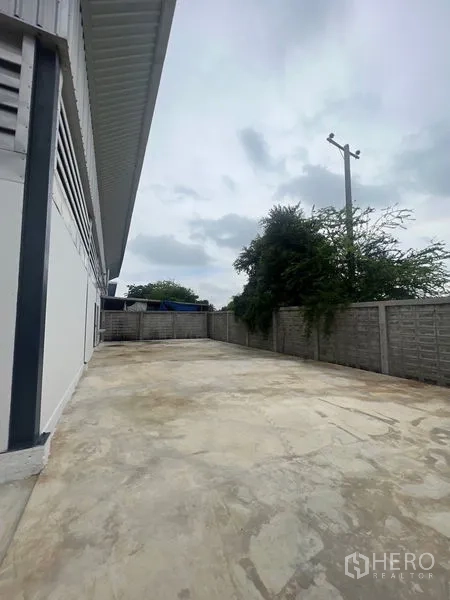 Warehouse for rent or sale in Sai Noi, Nonthaburi - Wide concrete yard along the warehouse with perimeter wall and trees.