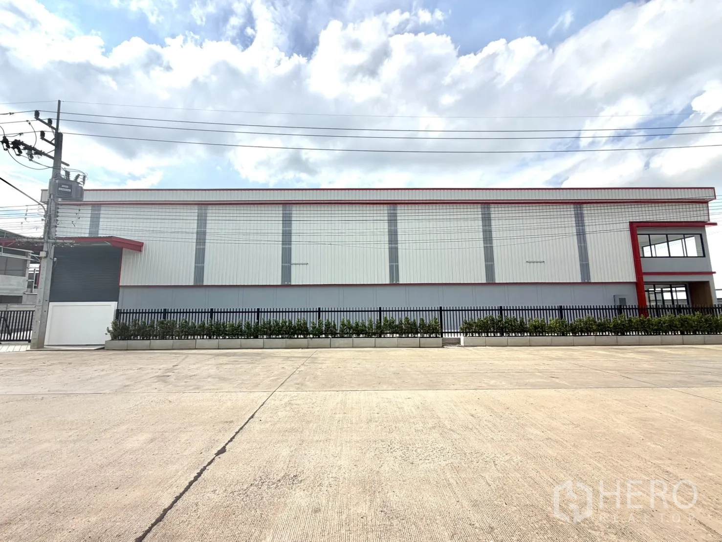 Factory for rent or sale in Lam Luk Ka, Pathum Thani - Long steel-clad warehouse facade behind a black fence along a wide road.