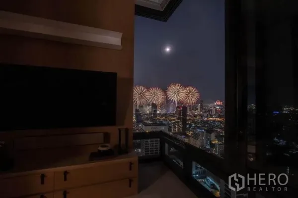 公寓 出租在Bang Rak, Bangkok - 夜间客厅视角，电视柜与阳台外的曼谷烟花与天际线。