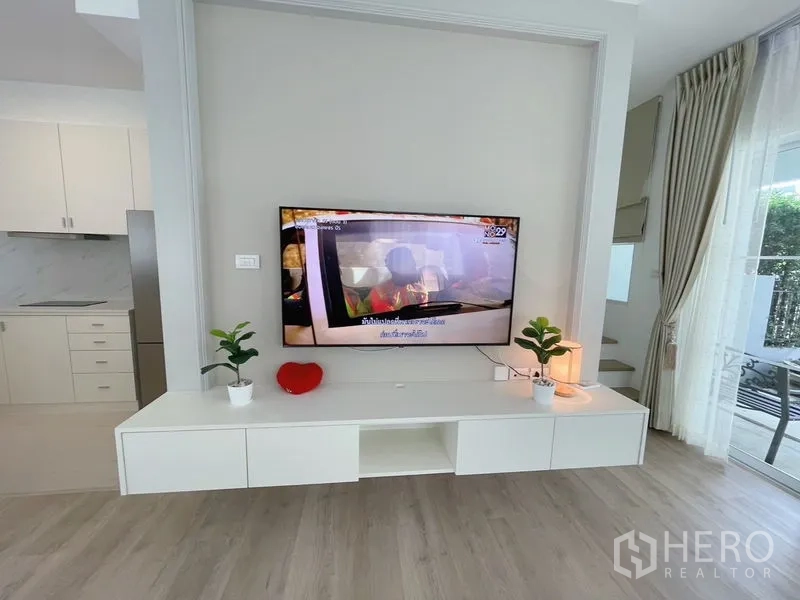 联排别墅 出租在Prawet, Bangkok - 电视特色墙配悬浮柜与绿植，旁连落地窗和楼梯。