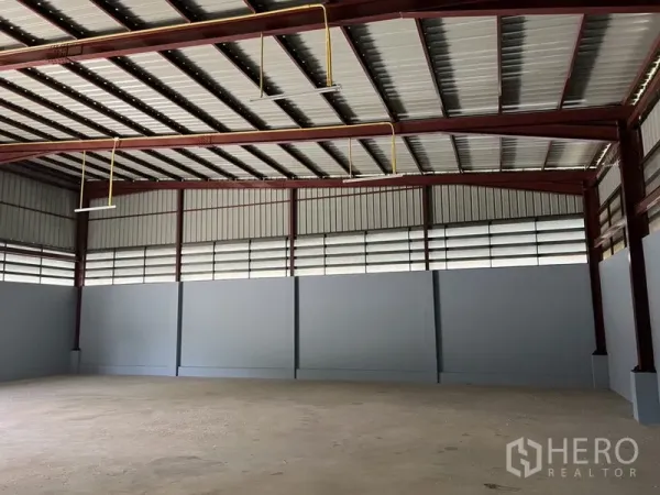 Warehouse for rent in Khlong Sam Wa, Bangkok - Clear-span storage hall with red steel beams, suspended lights, and louvered ventilation panels.