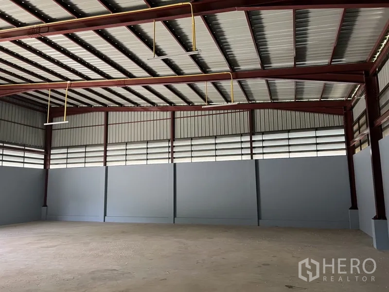 Warehouse for rent in Khlong Sam Wa, Bangkok - Clear-span storage hall with red steel beams, suspended lights, and louvered ventilation panels.