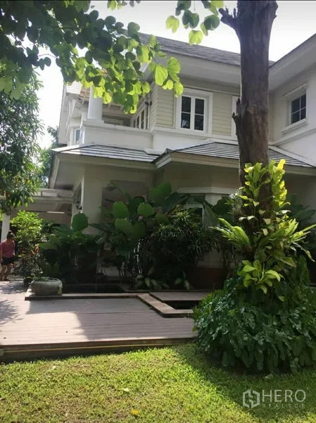 独立式住宅 出租在Phra Khanong, Bangkok - 白色两层住宅侧立面，木质露台与繁茂热带花园