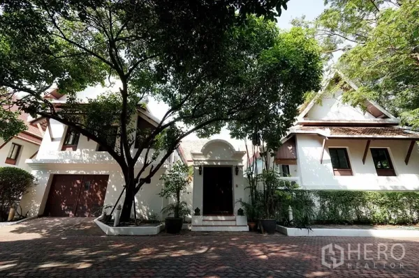 Detached House for rent in Watthana, Bangkok - Front exterior of a Thai-inspired detached house with garage, mature trees, and a brick-paved driveway.