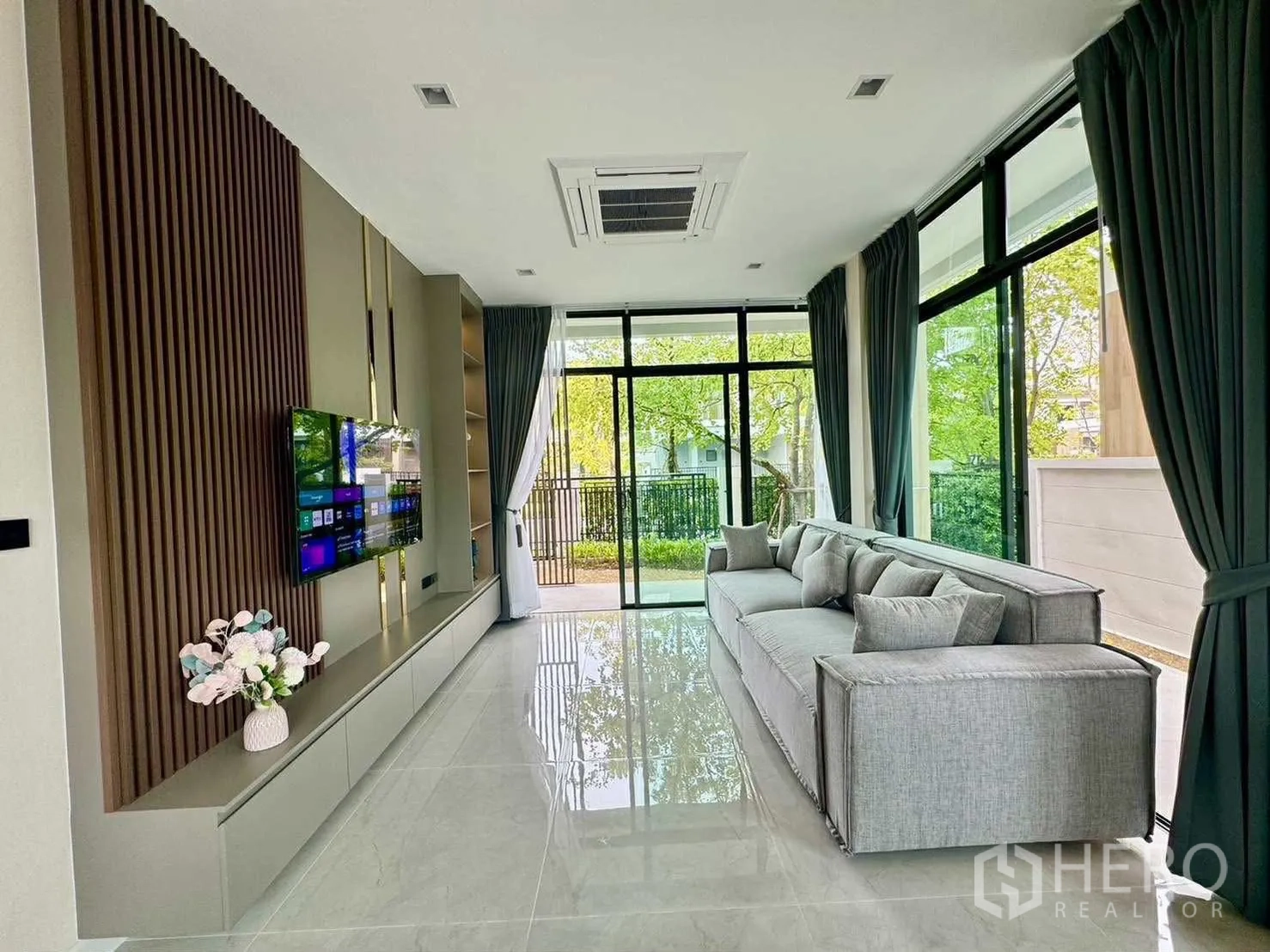 Detached House for rent in Bang Phli, Samut Prakan - Light-filled living room with long sofa, TV wall and large windows overlooking greenery.