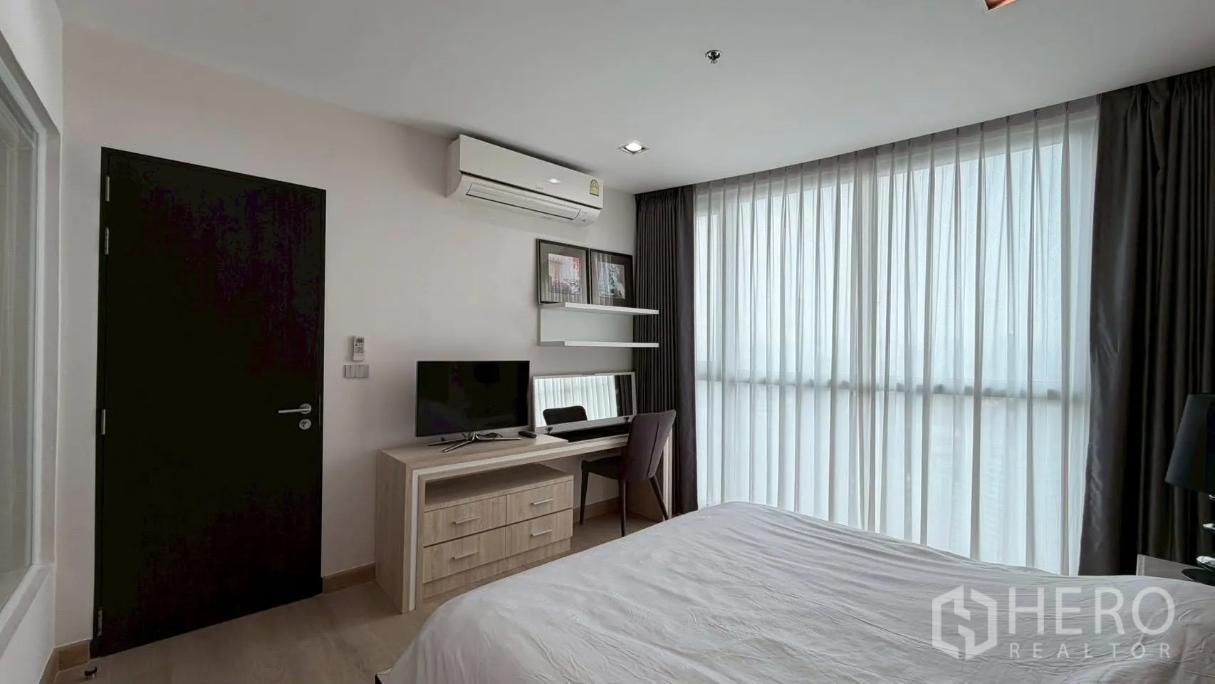 Condominium for rent or sale in Watthana, Bangkok - Bedroom view showing TV on a desk, wall shelves, air conditioner, and sheer curtains over tall windows.