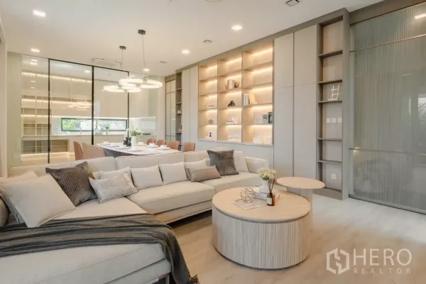 Detached House for rent in Saphan Sung, Bangkok - Open‑plan living and dining room with an L‑shaped sofa, round coffee tables, illuminated built‑in shelves, and a glass‑partition kitchen.