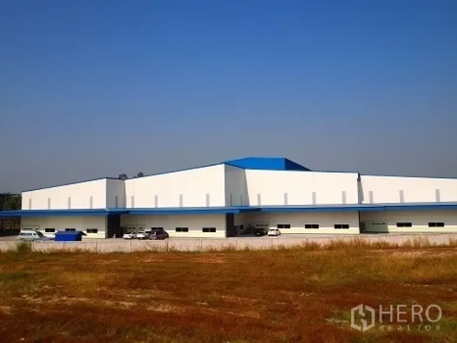 工厂 出租在Si Racha, Chon Buri - 从空地望向低矮的白色工厂外立面，蓝色屋顶与停车区清晰可见。