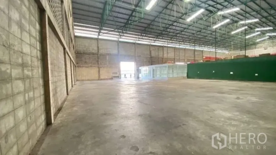 货栈 出租在Prawet, Bangkok - 高挑层高的仓库内景，钢结构屋顶、混凝土地面、宽卷闸出入口，侧边设玻璃办公室。