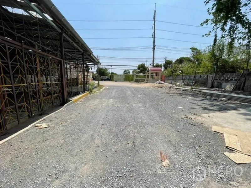 Warehouse for rent in Phra Samut Chedi, Samut Prakan - Wide gravel driveway and gated entrance with power lines at the industrial site.
