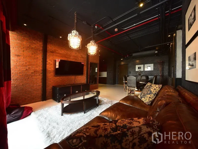 คอนโด ให้เช่าหรือขายในKhlong Toei, Bangkok - ห้องนั่งเล่นสไตล์ลอฟท์ ผนังอิฐ โซฟาหนัง โคมแขวน และทีวีติดผนังในคอนโดย่านสุขุมวิท