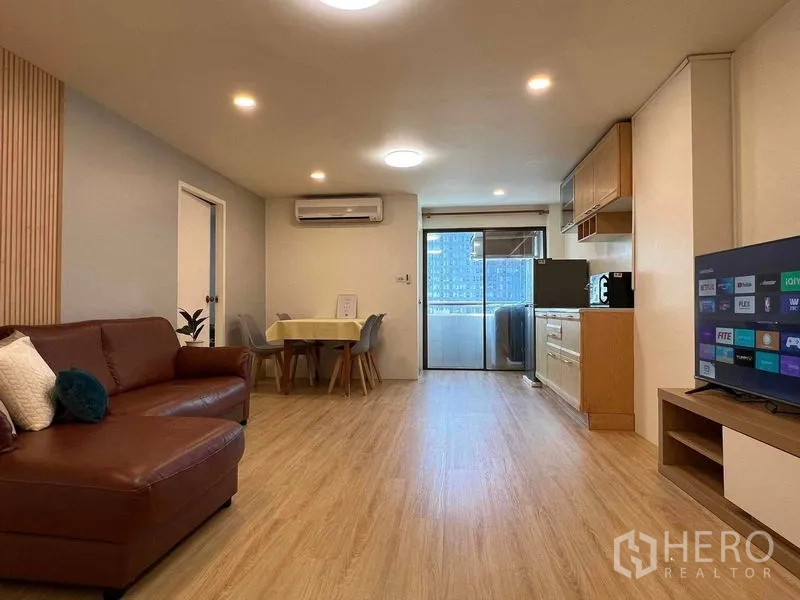 Condominium for rent in Ratchathewi, Bangkok - Open-plan living room with brown leather sofa, dining nook, kitchenette, and sliding glass door to the balcony.