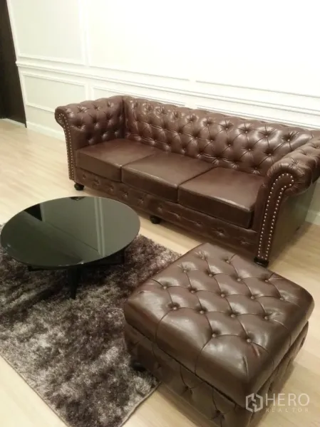 คอนโด ให้เช่าหรือขายในWatthana, Bangkok - โซฟาหนังสีน้ำตาลแบบหมอนลอนกับออตโตมาน โต๊ะกลางกระจกทรงกลมและพรมขนฟูบนพื้นไม้