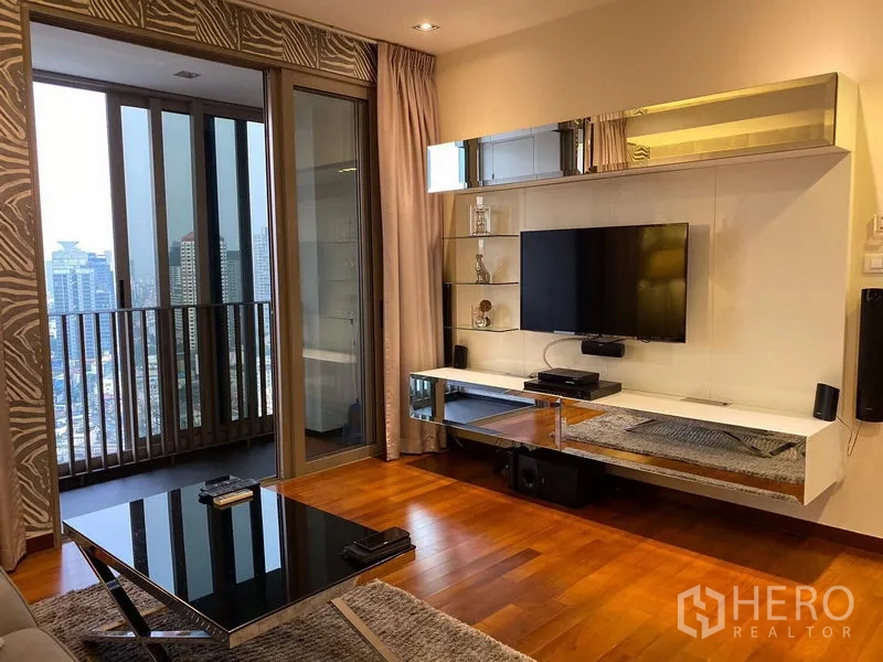 公寓 出租在Khlong Toei, Bangkok - 时尚电视墙与置物架，旁有玻璃门通往阳台，远眺城市天际线。